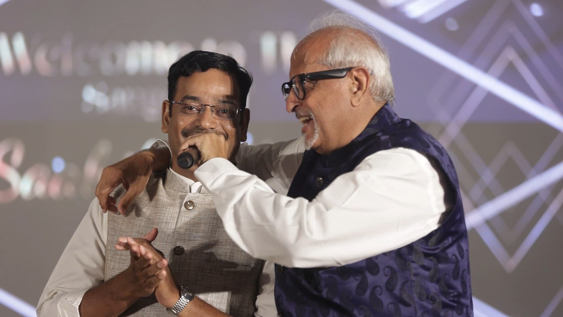 Ramesh Sadhwani on stage with his arm around a guest, holding the microphone to him as the two of them laugh