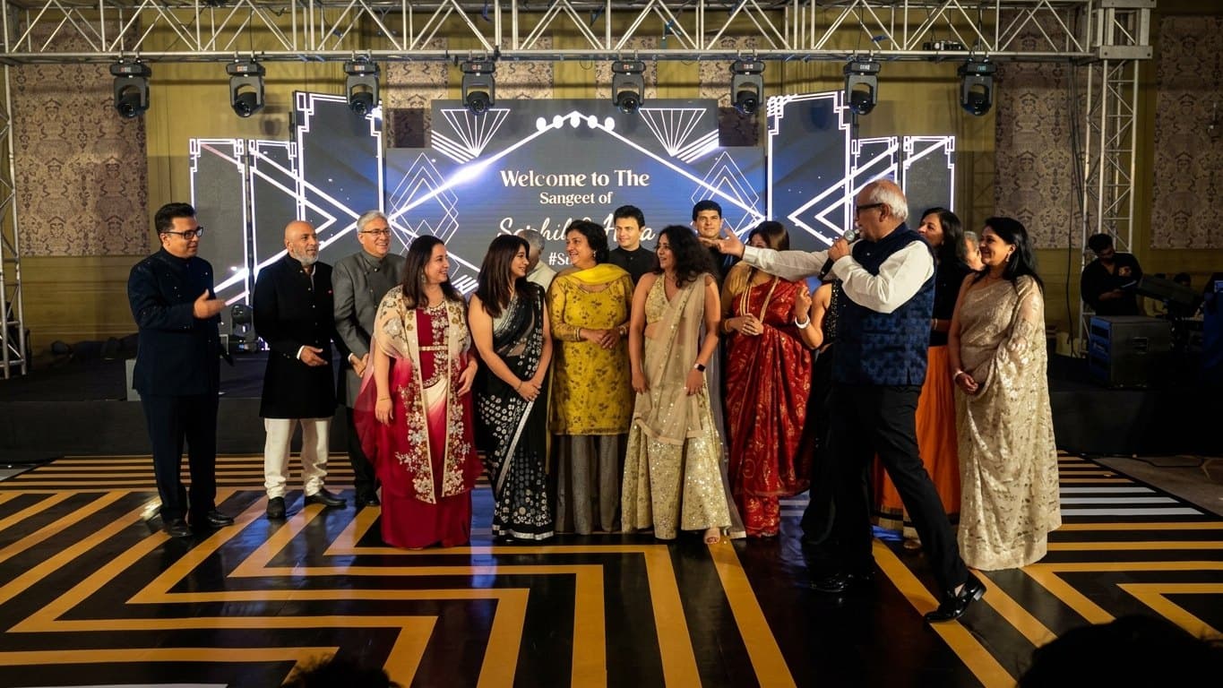 Ramesh Sadhwani at the microphone on a lit stage, directing a long line of family members standing together in front of a sangeet backdrop under a full lighting truss