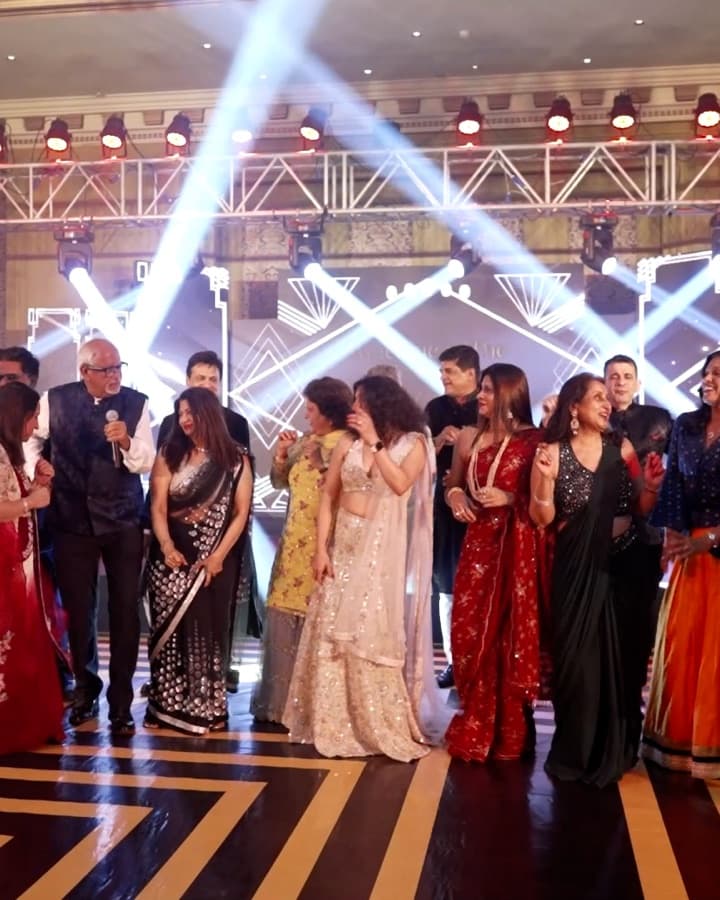 A full dance floor of guests singing and dancing together under stage lights, Ramesh Sadhwani among them with a microphone