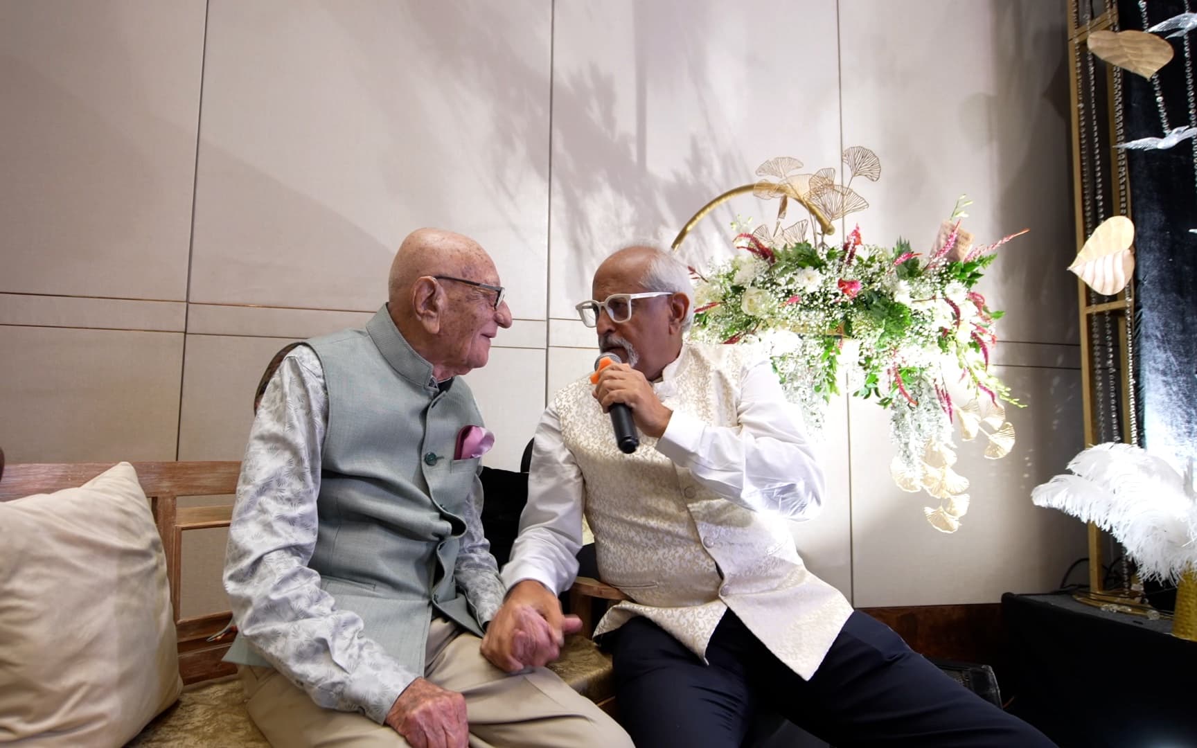 Ramesh Sadhwani seated beside an elderly guest of honour, holding a microphone for him as the two talk
