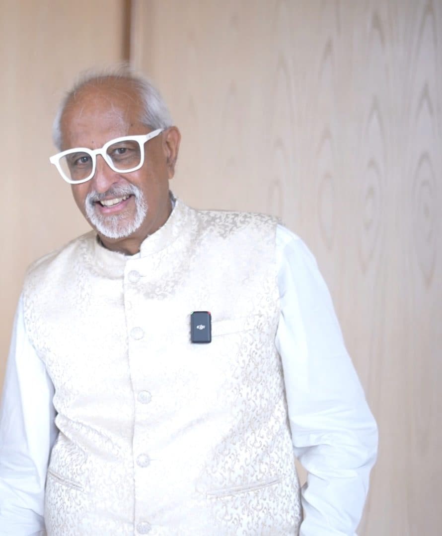 Ramesh Sadhwani in a white kurta and cream jacket, smiling mid-conversation with a lapel microphone clipped on, in a hotel foyer